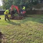 Two men wearing bright green construction worker uniforms plow the dirt in a residential backyard.